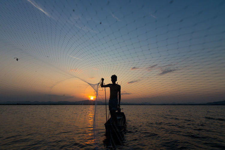Silhouette of man throwing fishing net while standing in boat on sea against sky during sunset