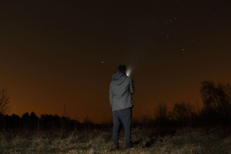 Rear view of man with flashlight standing on field against sky at night