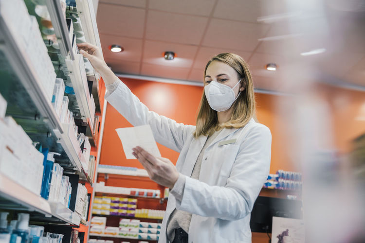 Female pharmacist wearing protective face mask while working in chemist shop