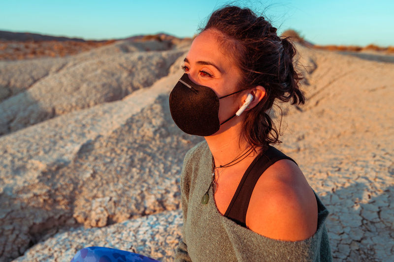 Side view of confident young sportswoman in black protective mask listening to music with wireless earbuds while recreating during training in rough desert badlands looking away