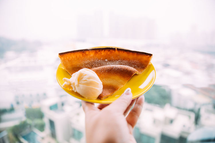 Cropped image of woman hand holding ice cream with pancake in plate against cityscape