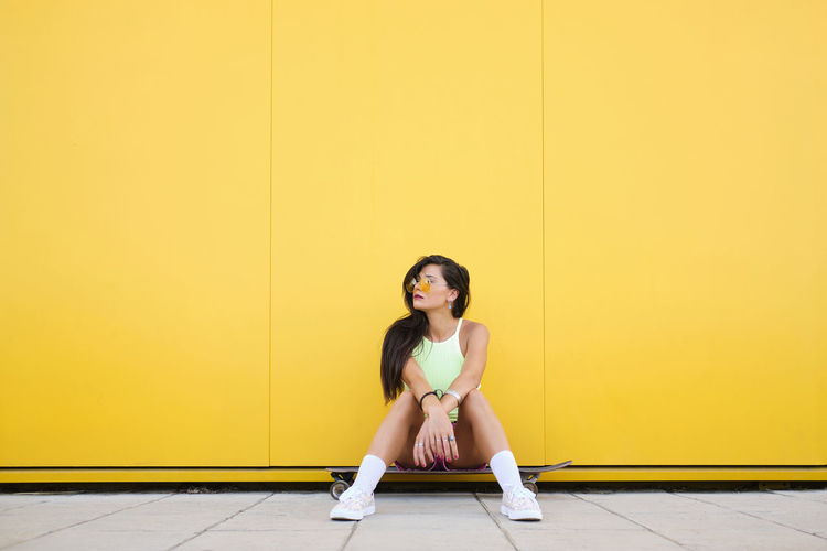 Portrait of beautiful girl sitting alone on longboard in front of yellow wall
