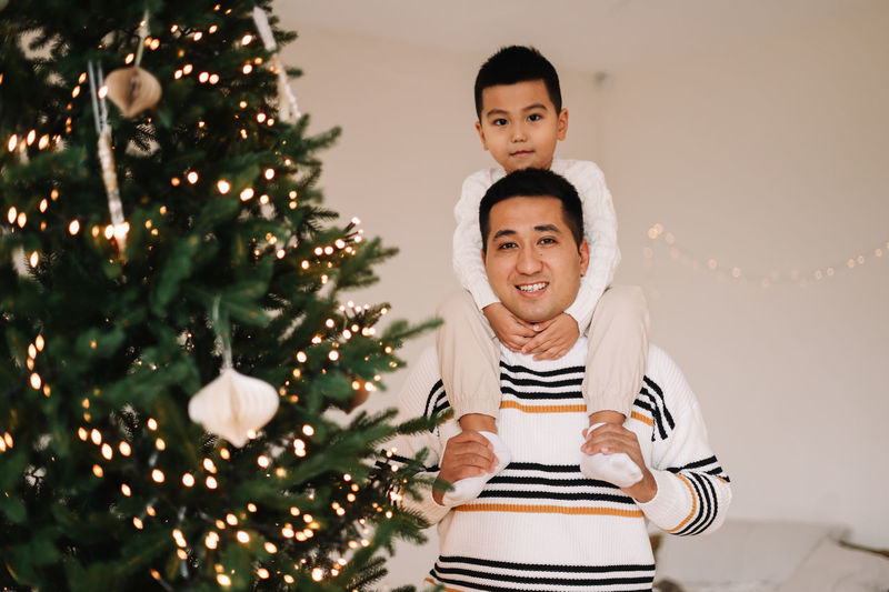 Chinese multiracial dad and little son in sweaters decorate the christmas tree at home on holiday