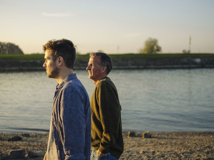 Father and son looking away while spending leisure time by river at evening