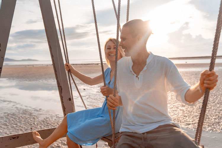 
close-up of a man and a woman riding on a swing against the rising sun on the seashore