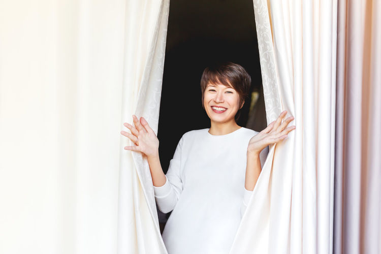 Wide smiling beautiful asian woman opens curtains on window. woman among textile folds of drapery.