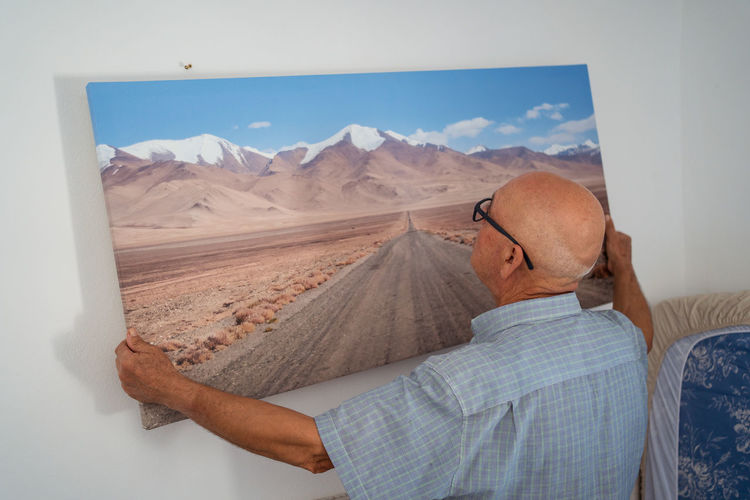 Back view of senior bald male in eyeglasses and checkered shirt hanging artwork of road and mountains on wall at home