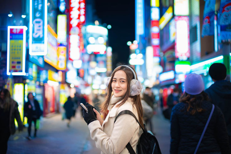 Portrait of smiling young woman using phone on illuminated street at night