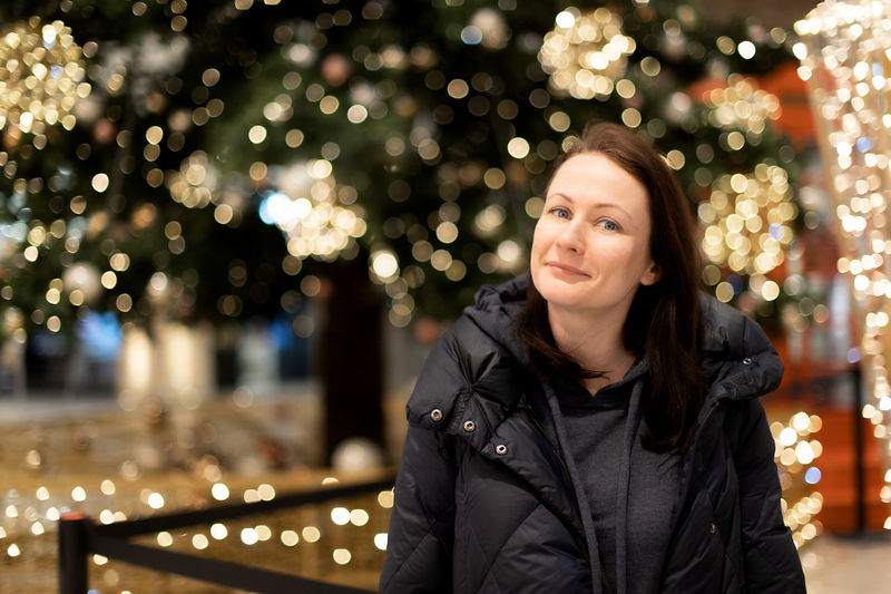 Portrait of young woman standing against illuminated christmas tree at night