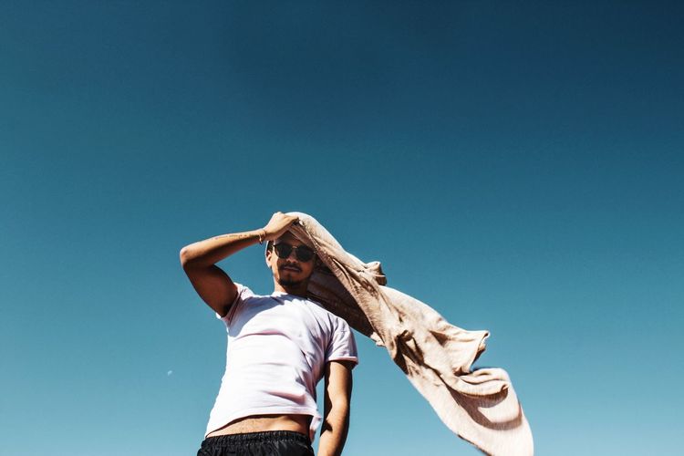 Low angle portrait of man holding jacket while standing against clear blue sky