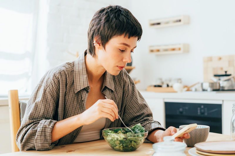 A young short-haired brunette  in the kitchen eating and browsing social networks on her phone.