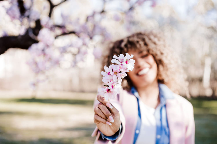 Happy hispanic woman with afro hair in spring among pink blossom flowers. selective focus on flowers