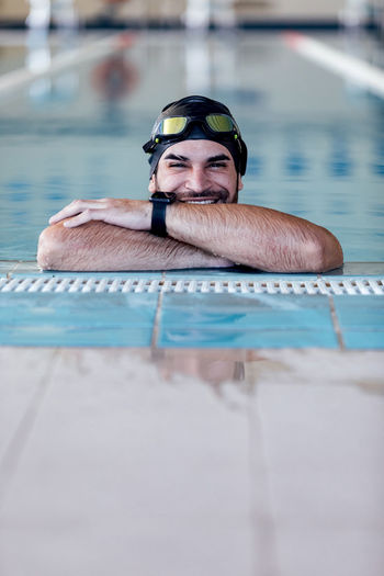 Cheerful sportsman in swimming cap and goggles looking at camera from pool on blurred background