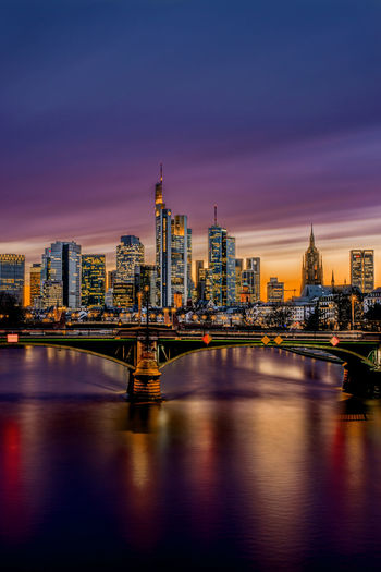 Illuminated bridge over river by buildings against sky at sunset in frankfurt, germany. 