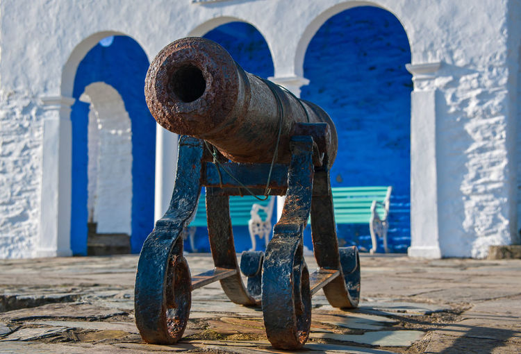 Old rusty metallic cannon against blue walls and building