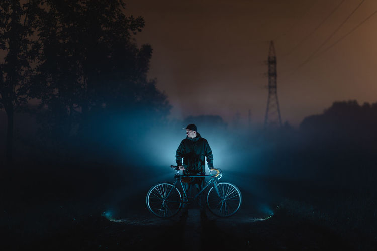 Man with bicycle against illuminated light standing on road at night