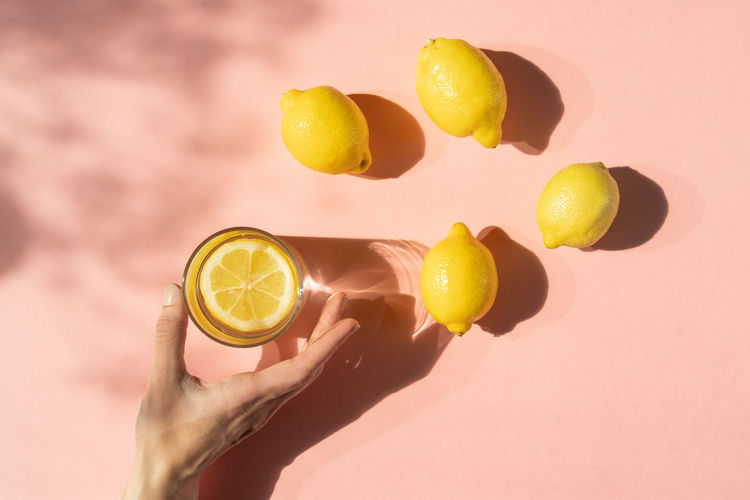 Close-up of hand with drinking glass and lemons on pink background