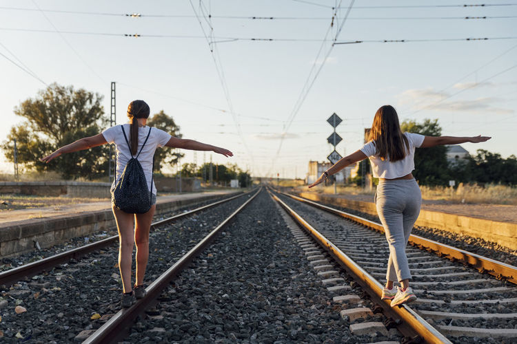 Female friends with arms outstretched walking on railroad track against sky at sunset