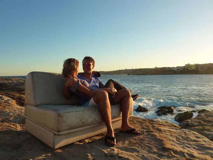 Couple sitting on sofa at beach against clear sky