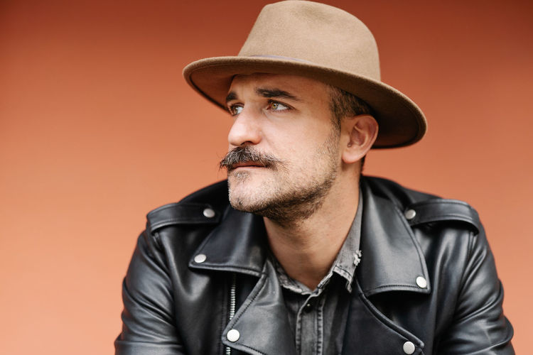 Trendy thoughtful male in hat and leather jacket on beige background looking away