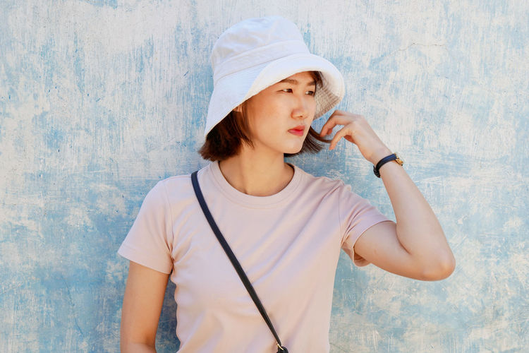 Beautiful woman wearing hat looking away while standing against wall