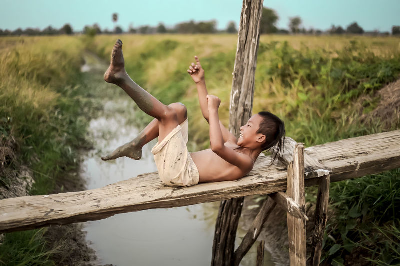 Happy shirtless boy lying on wood over canal at farm
