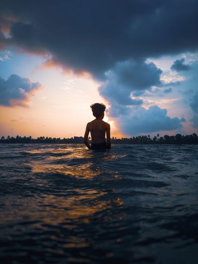 Rear view of shirtless boy in sea against sky during sunset