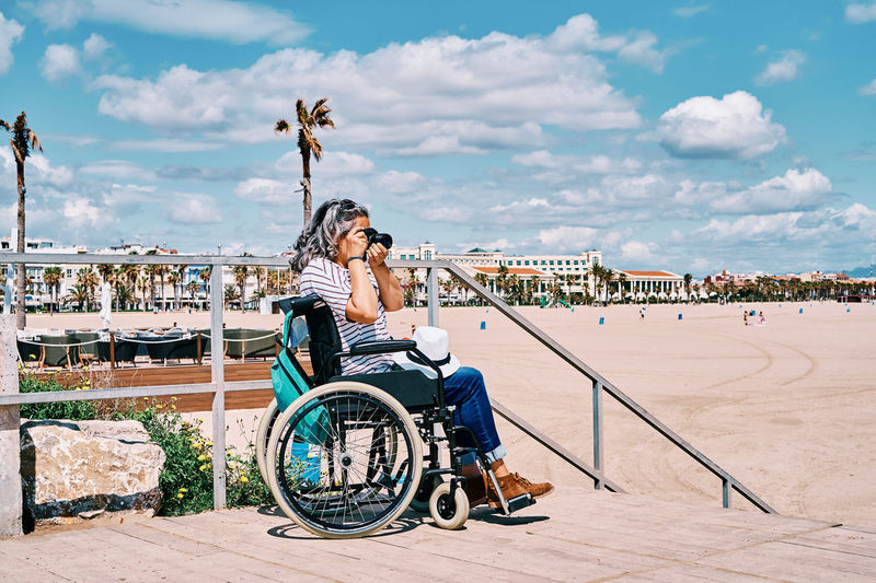 Side view of unrecognizable gray haired disabled woman in wheelchair taking pictures on photo camera while spending summer day on beach