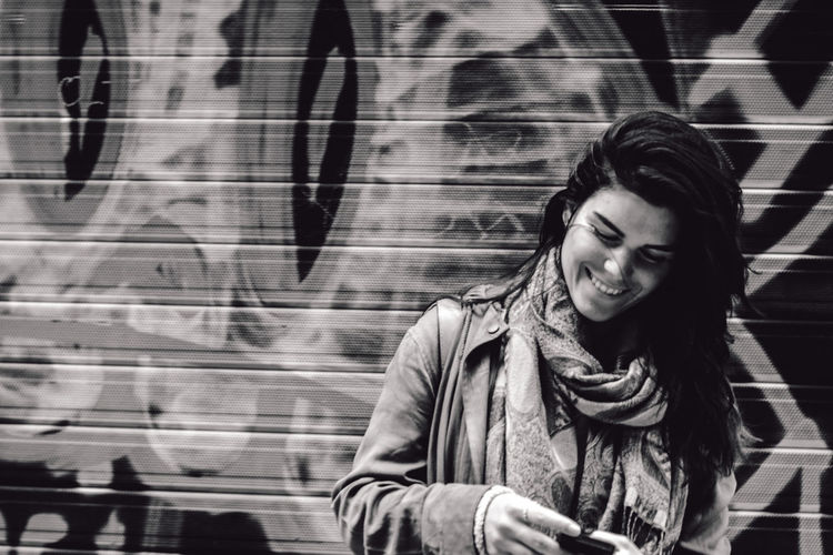 Close-up of happy woman using phone while standing by graffiti wall