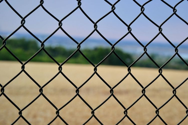 Full frame shot of chainlink fence, with a wheat field and blue sky in the background