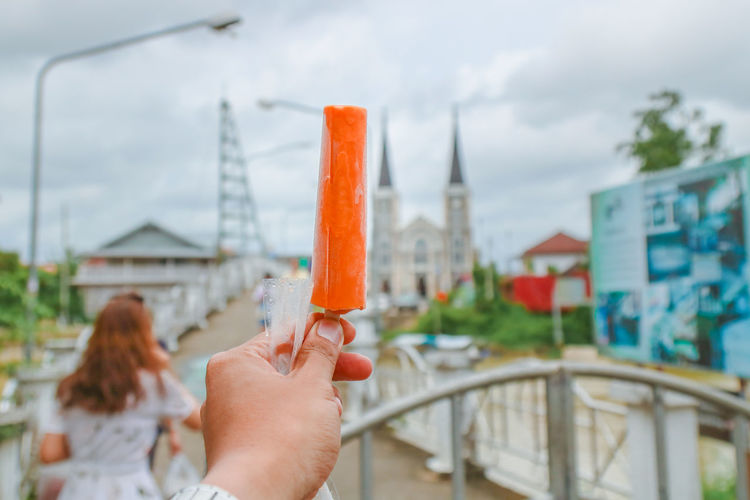 Cropped hand holding popsicle against sky in city