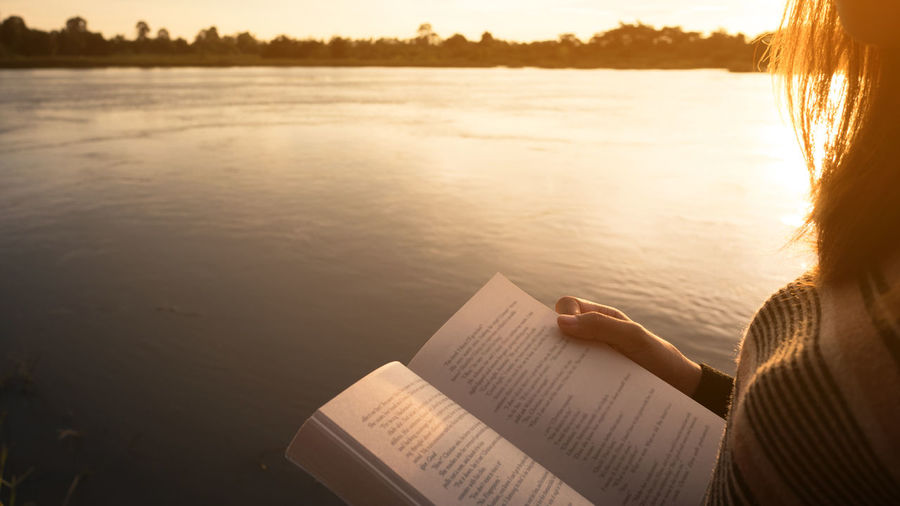Rear view of woman reading book by lake during sunset