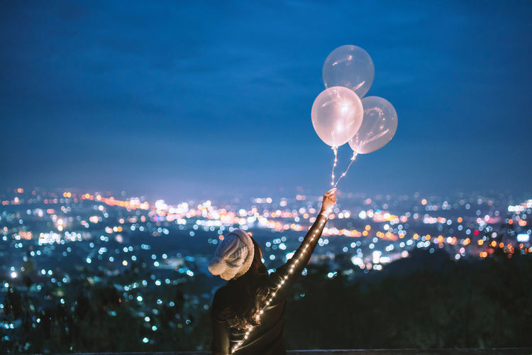 Rear view of woman with arms raised holding balloons while standing against cityscape at night