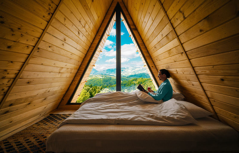 Man reading book and drinking wine in bed in a-frame wooden cabin at mountain