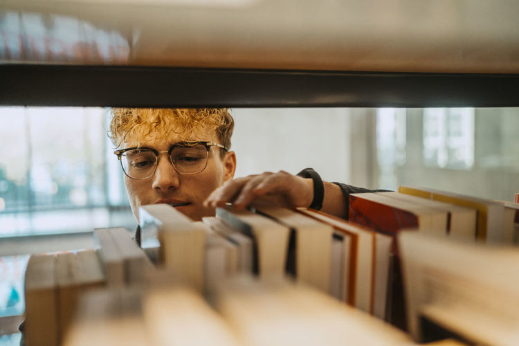 Young man wearing eyeglasses searching book in library at university