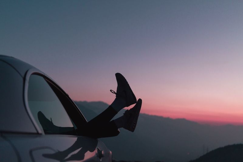 Low section of woman peeking from car window against sky during sunset