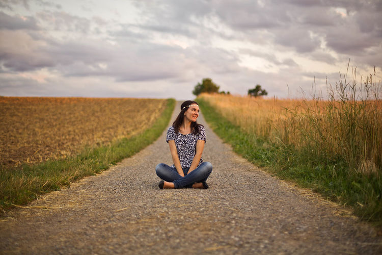 Smiling woman sitting on road amidst field against cloudy sky