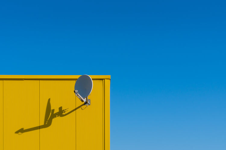 Low angle view of satellite dish at yellow wall against clear blue sky