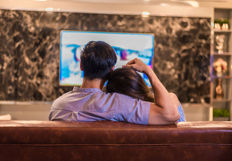 Rear view of couple watching television while sitting on sofa at home