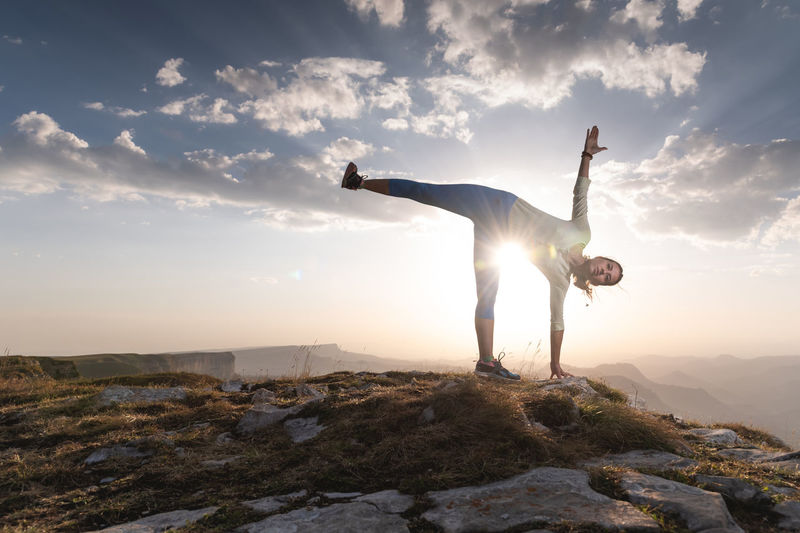 Young woman doing beautiful yoga, sunset or sunrise in mountains over blue sky and clouds against
