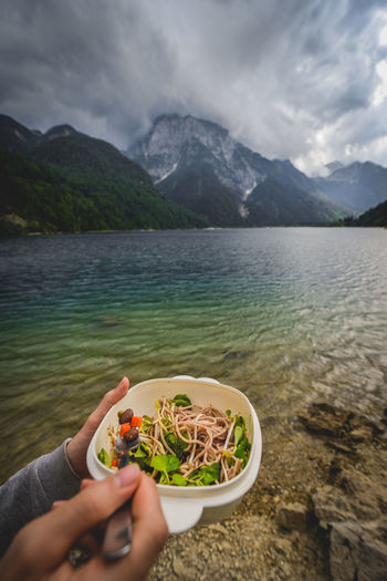 Cropped hands having noodles at lakeshore against cloudy sky
