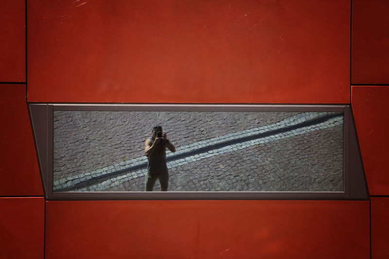 Reflection of man photographing while standing on footpath in mirror over building