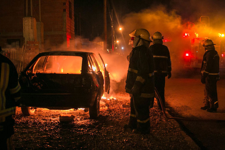 Firefighters standing by burning car at night