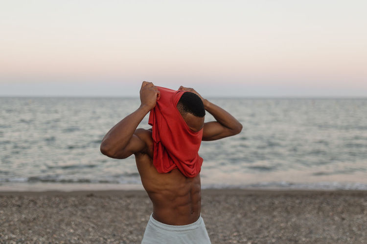 Cheerful african american male runner putting on t shirt while resting on seashore and smiling brightly