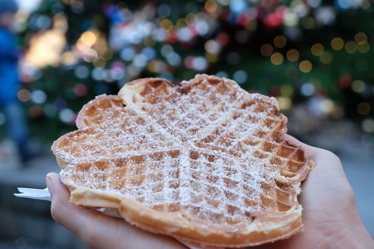 Two hands holding brown crispy waffle with icing sugar. colourful bokeh christmas balls and market.
