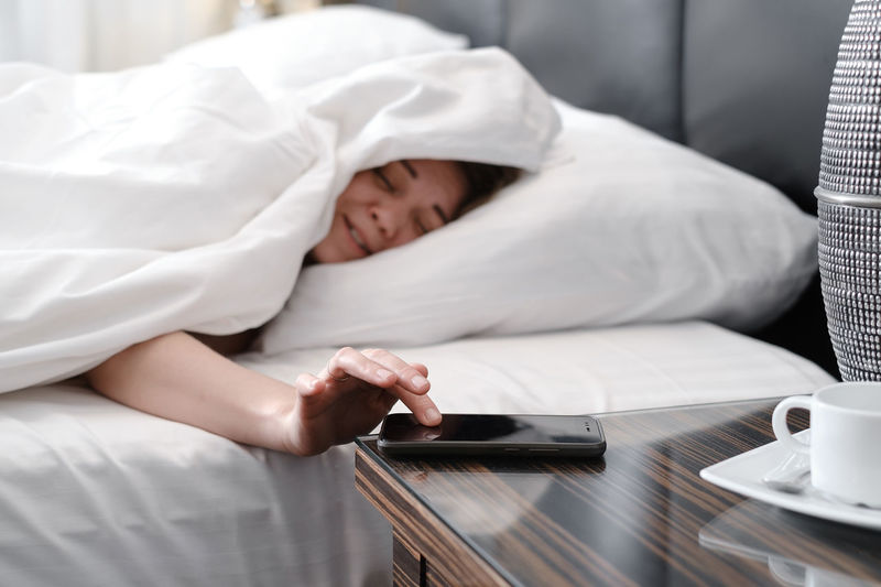 Sleepy woman covered with blanket in the morning in bed turning off alarm clock on cell phone.