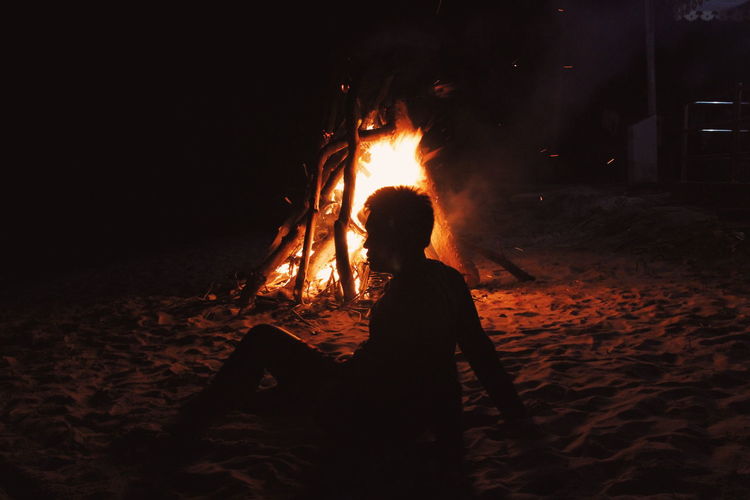 Silhouette man sitting by burning bonfire at campsite