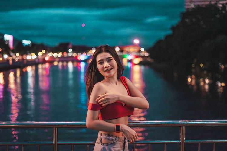 Portrait of smiling young woman standing on bridge over river at night