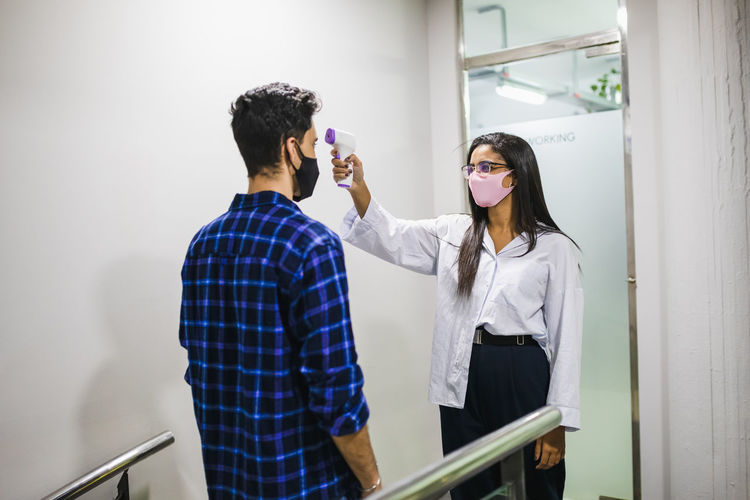 Businesswoman measuring temperature of happy colleague using digital thermometer on yellow background