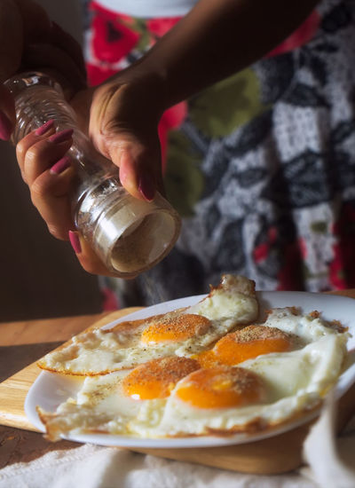 Cropped hands of woman sprinkling pepper on fried eggs at home